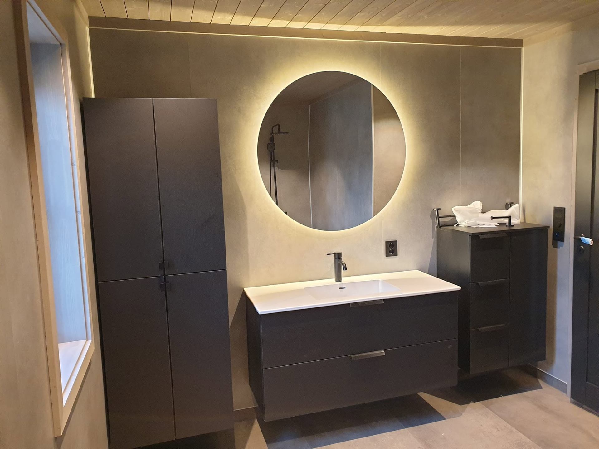 Moderne bad med mikrosement, sort innredning og rundt speil med baklys – levert av Byggservice 1 AS